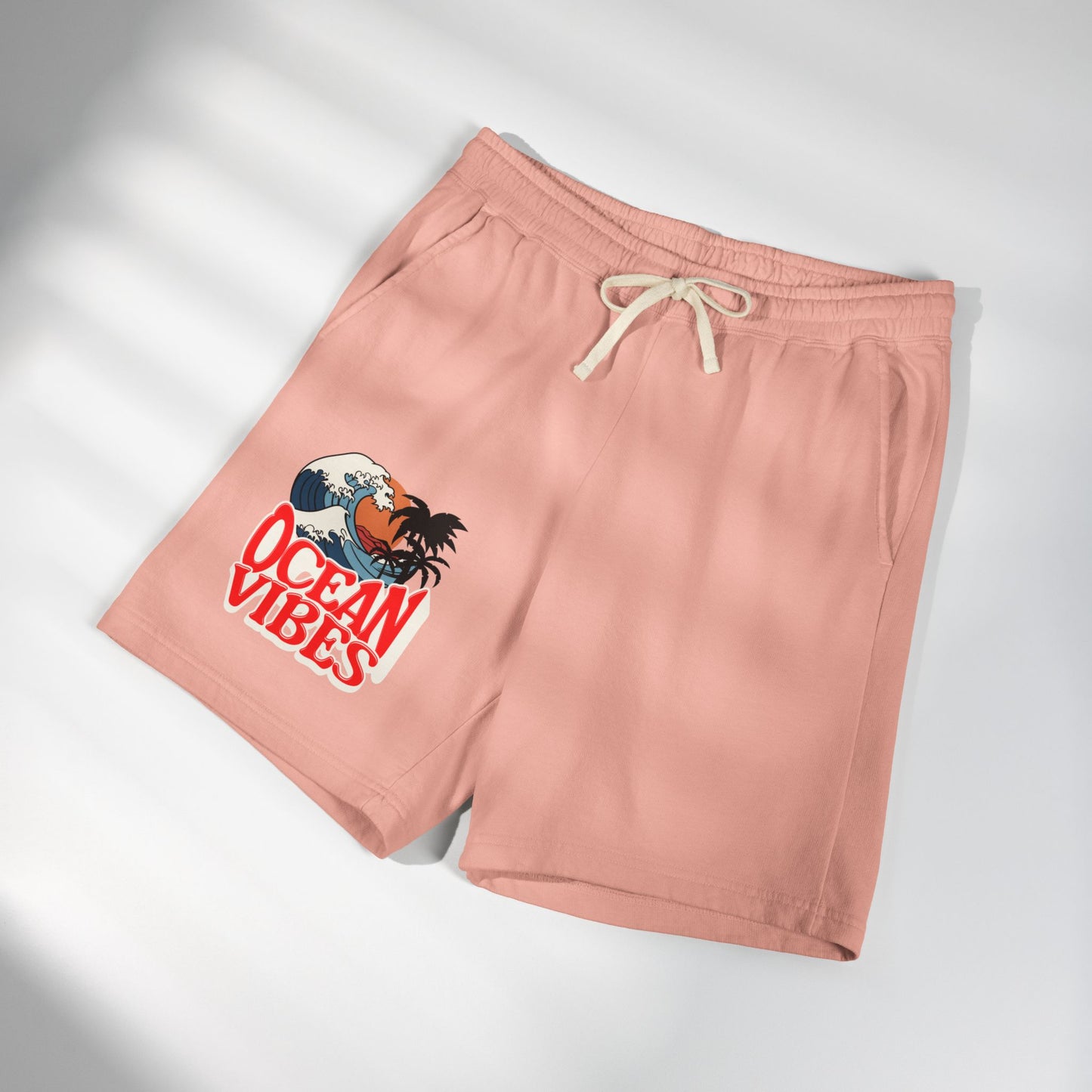 A pair of dusty pink athletic shorts with a cream drawstring, featuring a graphic print of a Hawaiian character in a wave with the text 'OCEAN VIBES' on the left leg, displayed on a light gray background with soft diagonal shadows. Unique Lightweight Fleece Sweat Shorts in Ocean Vibes Graphic Shop Lightweight Fleece Sweat Shorts in Ocean Vibes for breezy comfort, relaxed style, and an easygoing summer-ready look all day. Refresh your casual rotation with Lightweight Fleece Sweat Shorts in Ocean Vibes.