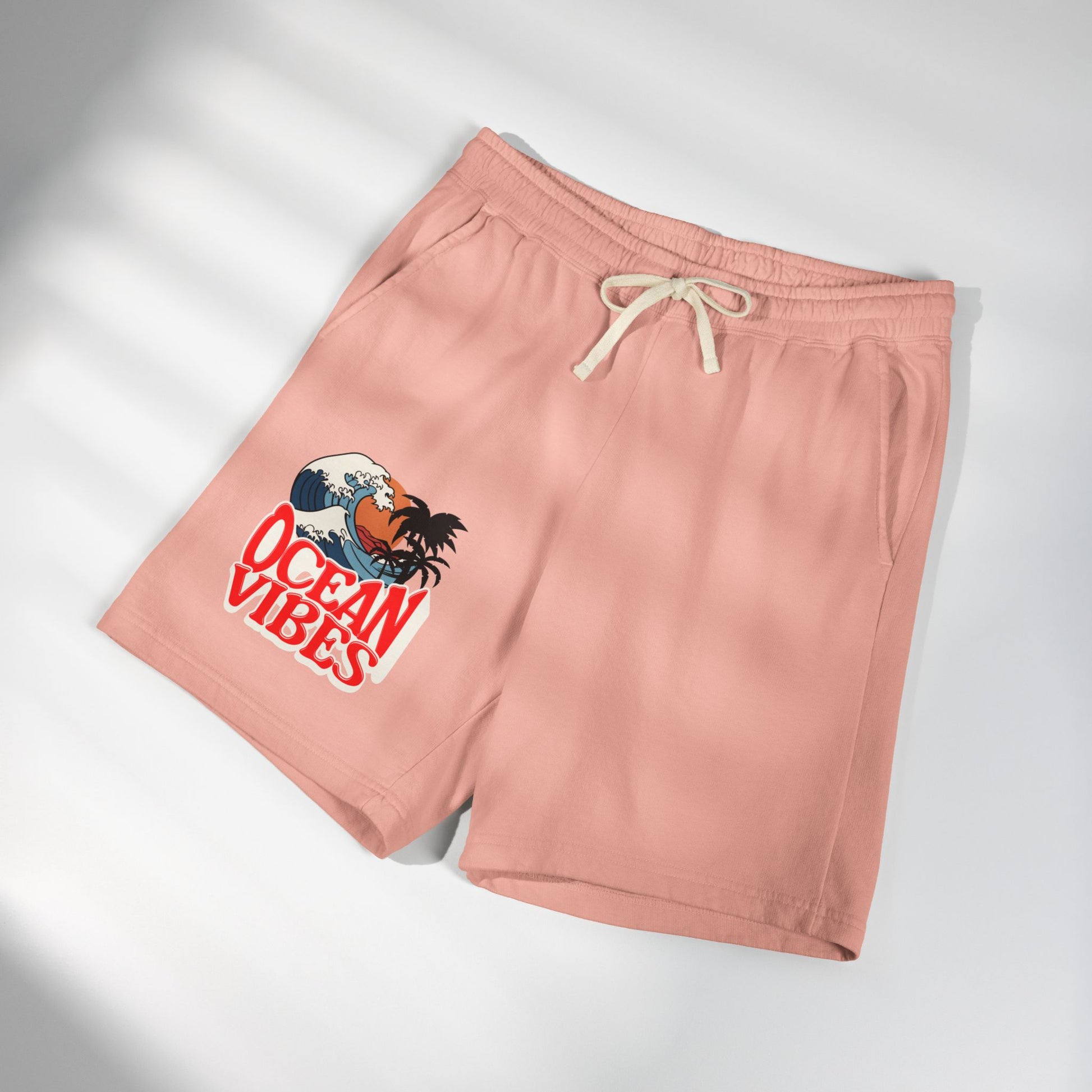 A pair of dusty pink athletic shorts with a cream drawstring, featuring a graphic print of a Hawaiian character in a wave with the text 'OCEAN VIBES' on the left leg, displayed on a light gray background with soft diagonal shadows. Unique Lightweight Fleece Sweat Shorts in Ocean Vibes Graphic Shop Lightweight Fleece Sweat Shorts in Ocean Vibes for breezy comfort, relaxed style, and an easygoing summer-ready look all day. Refresh your casual rotation with Lightweight Fleece Sweat Shorts in Ocean Vibes.