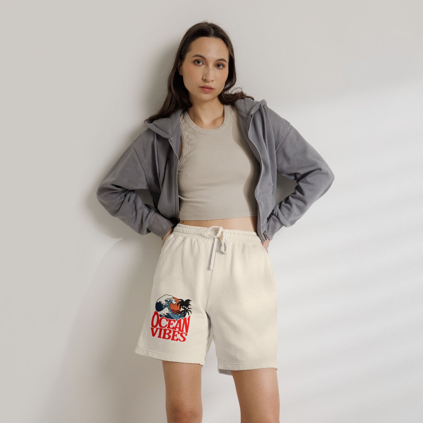 A woman stands against a light gray wall wearing cream-colored sweat shorts with a red and blue 'Ocean Vibes' graphic on the left leg, a beige ribbed crop top, and a gray zip-up hoodie. Unique Lightweight Fleece Sweat Shorts in Ocean Vibes Graphic Shop Lightweight Fleece Sweat Shorts in Ocean Vibes for breezy comfort, relaxed style, and an easygoing summer-ready look all day. Refresh your casual rotation with Lightweight Fleece Sweat Shorts in Ocean Vibes—soft, versatile, and ready for sunny days.