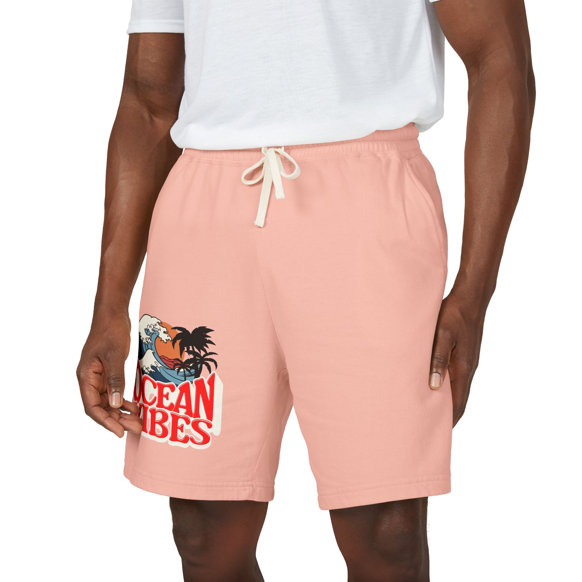 A person wearing peach-pink drawstring shorts with a graphic print on the left leg that reads 'Ocean Vibes' with a tropical wave and palm design, paired with a white t-shirt against a plain white background.
