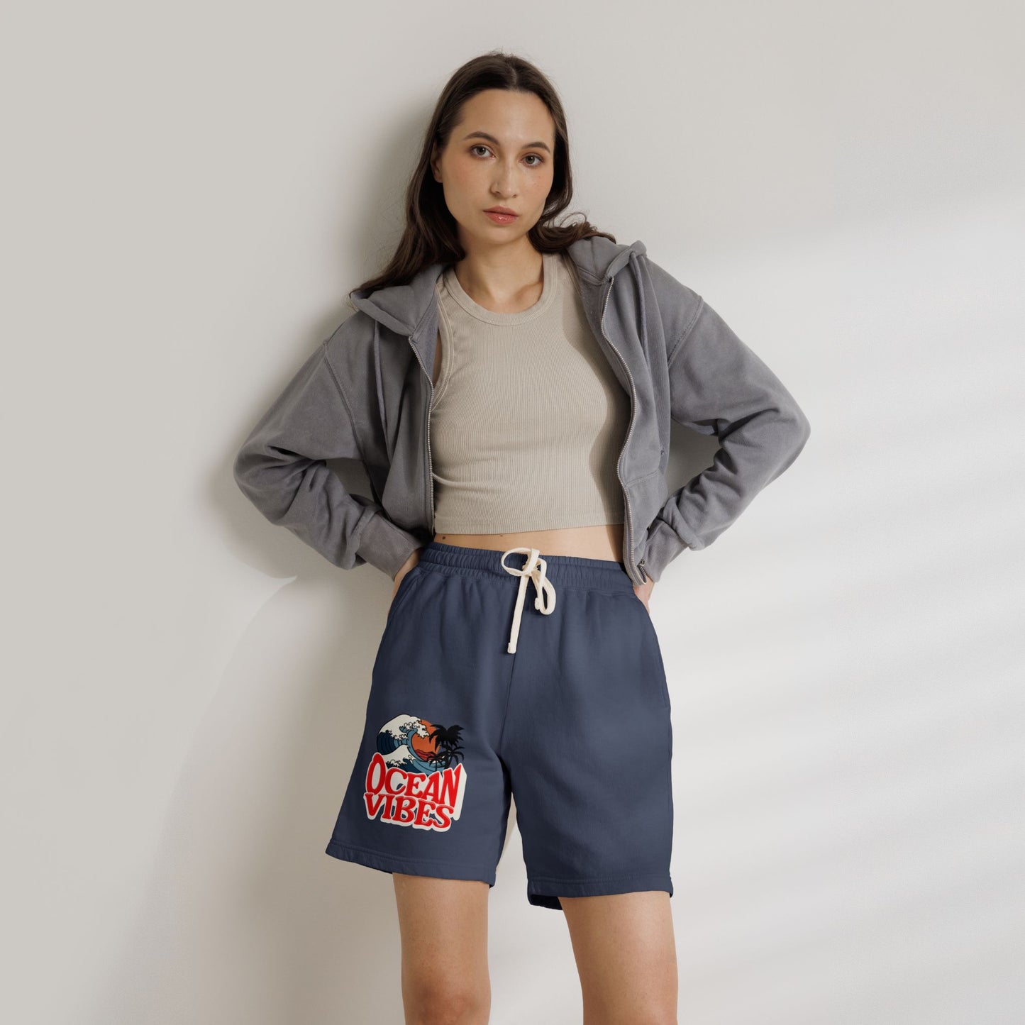 A woman modeling navy blue drawstring shorts with a red and white 'Ocean Vibes' graphic on the left leg, styled with a beige crop top and a gray zip-up hoodie against a light gray wall.