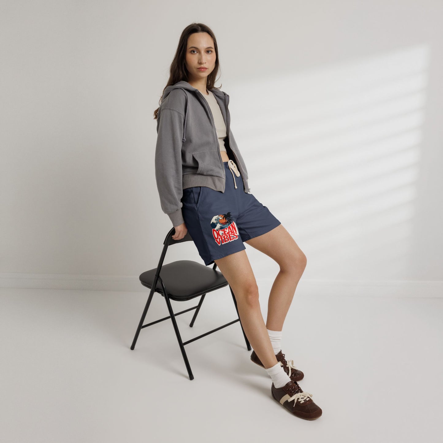 A woman models navy blue drawstring shorts with a colorful graphic print on the left leg, styled with a gray cropped hoodie, white t-shirt, white socks, and brown loafers, while leaning on a black folding chair against a white wall with soft window shadows.