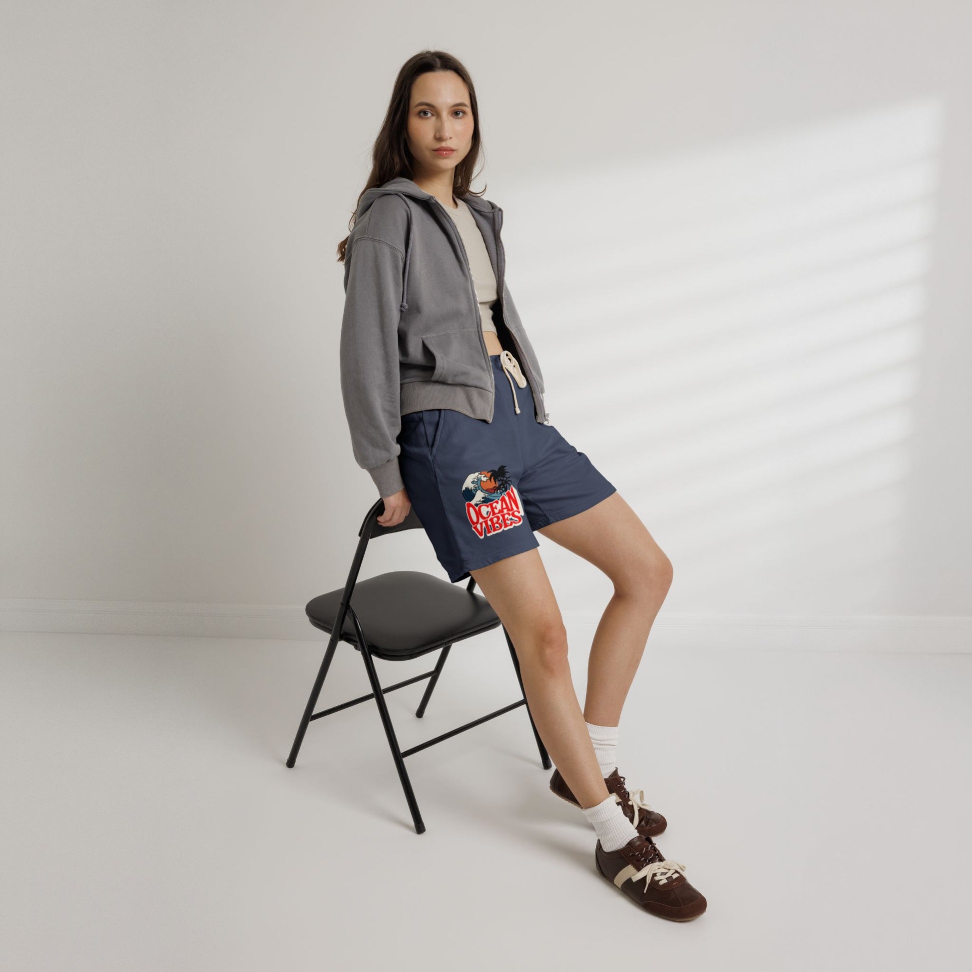 A woman models navy blue drawstring shorts with a colorful graphic print on the left leg, styled with a gray cropped hoodie, white t-shirt, white socks, and brown loafers, while leaning on a black folding chair against a white wall with soft window shadows.