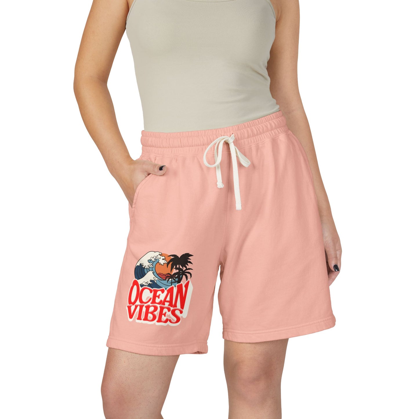 A model wearing light peach-pink drawstring shorts with a 'Ocean Vibes' graphic on the left leg, paired with a beige ribbed tank top against a plain white background.