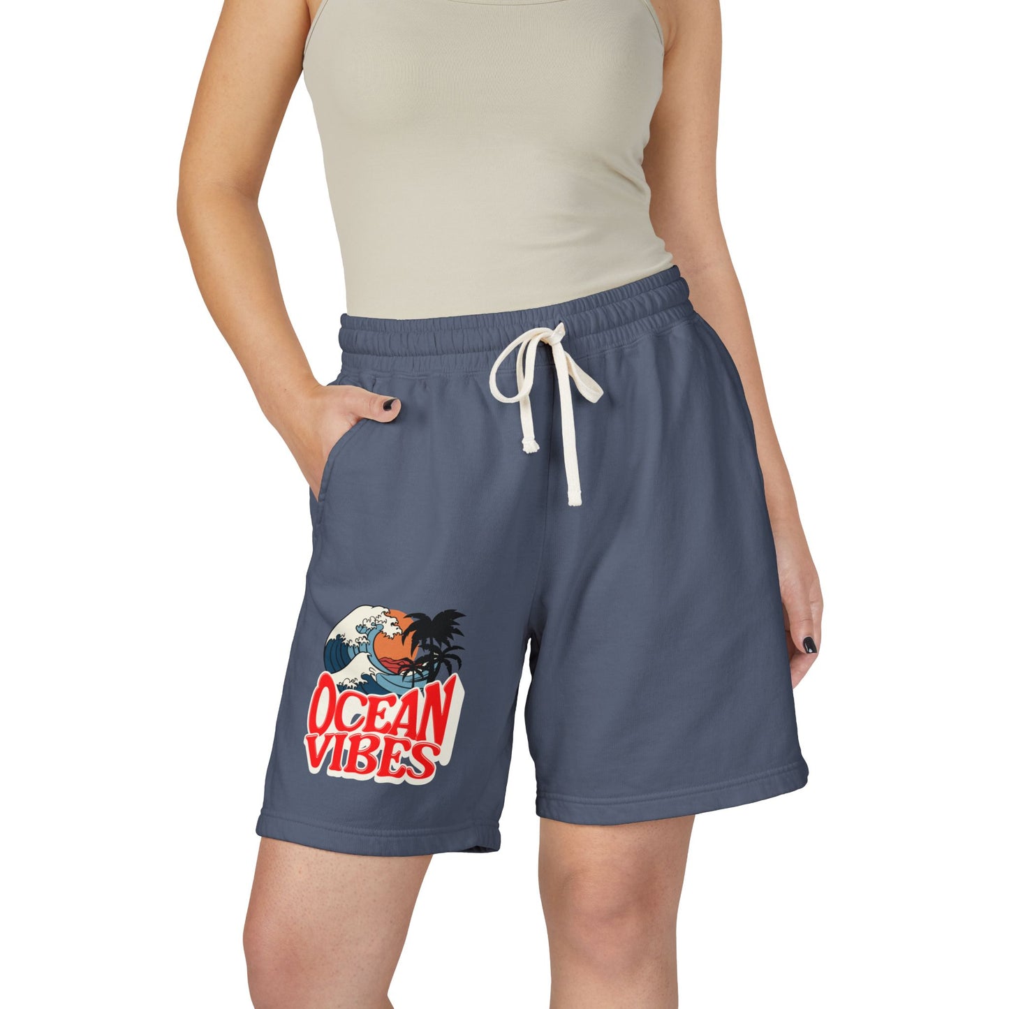 A model wearing slate blue drawstring shorts with a 'Ocean Vibes' graphic on the left leg, paired with a beige ribbed tank top against a plain white background.