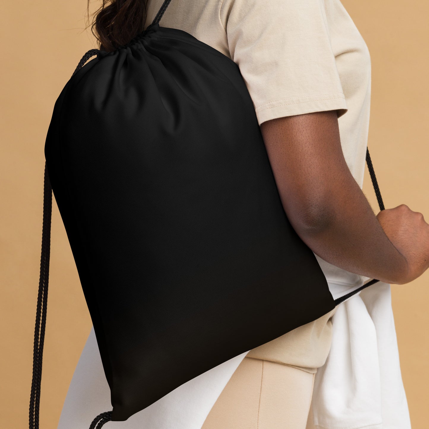 A person wearing a beige t-shirt and white shorts carries a black drawstring backpack over their shoulder. The backpack features a patriotic red, white, and blue candy cane graphic on the front, set against a solid beige background. Bold Minimalist Solid Black Lightweight Drawstring Bag