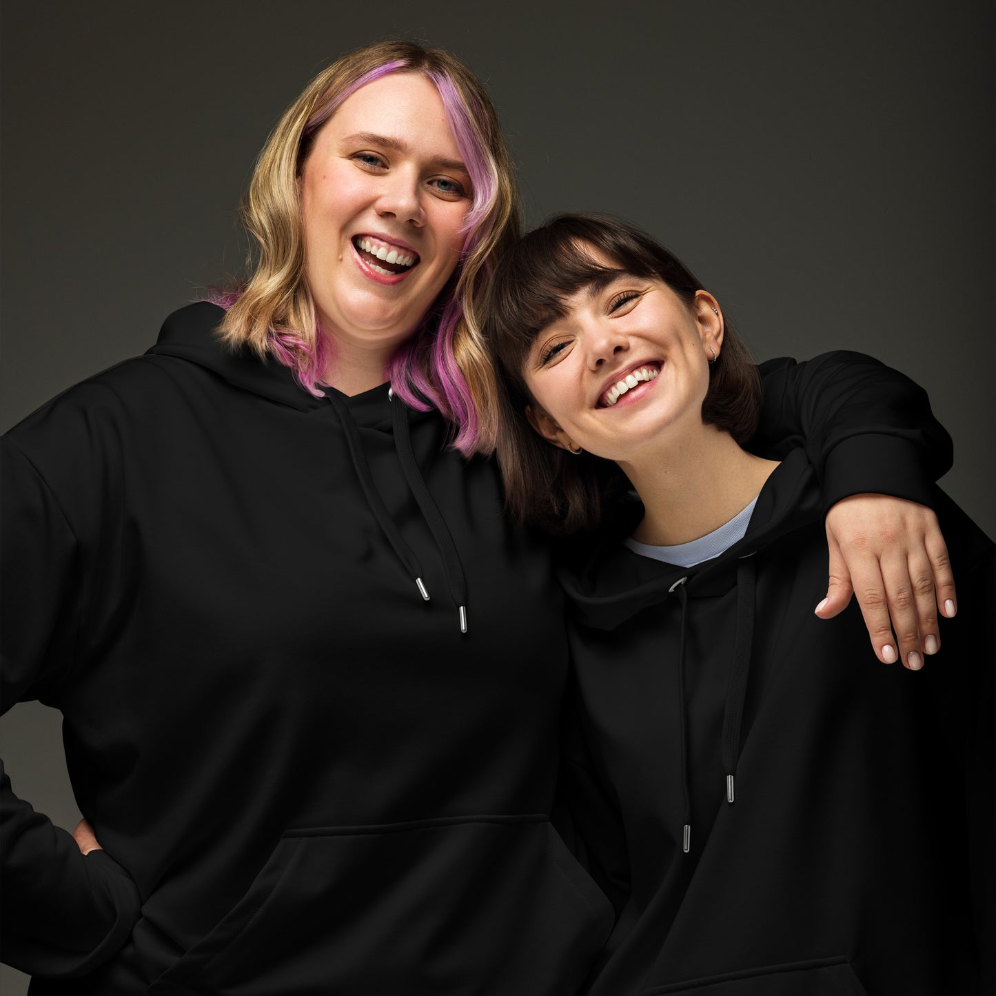 Two women smiling and posing together in matching black hoodies against a dark gray studio background. Bold Minimalist Solid Plain Dark Black Hoodie for Casual Wear