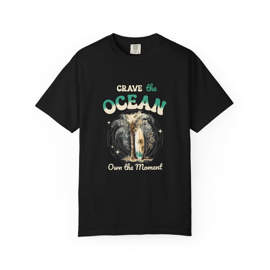 A black short-sleeve crewneck t-shirt with a graphic on the front that reads 'Crave the OCEAN Own the Moment' in stylized teal and white lettering, surrounding a central illustration of a surfer on a board. Crave the Ocean Own the Moment Graphic T-Shirt for Casual Wear