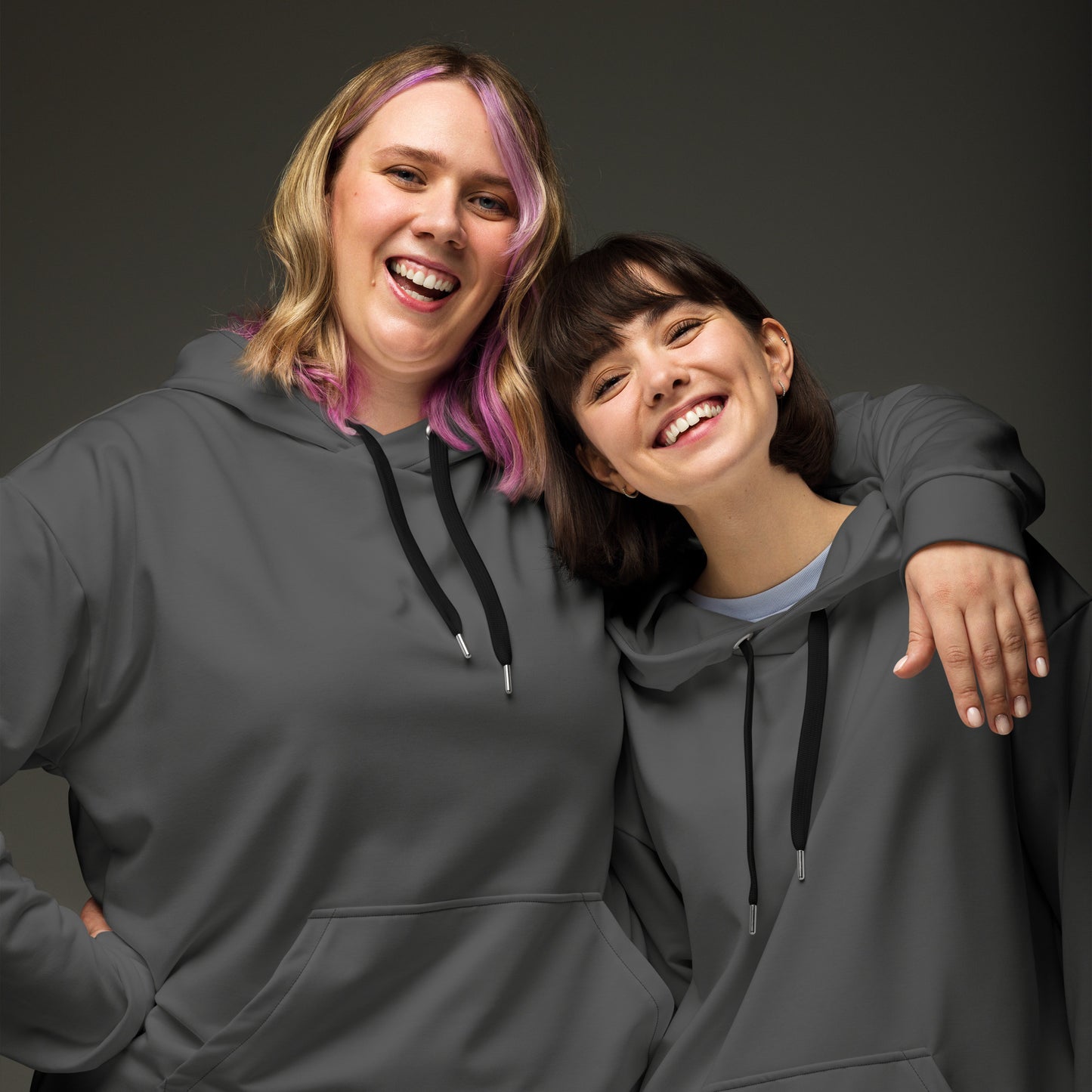 Two women smiling and posing together while wearing matching dark gray hoodies with black drawstrings, photographed against a dark gray studio background. Minimalist Comfortable Plain Gray Hoodie for Relaxed Wear
