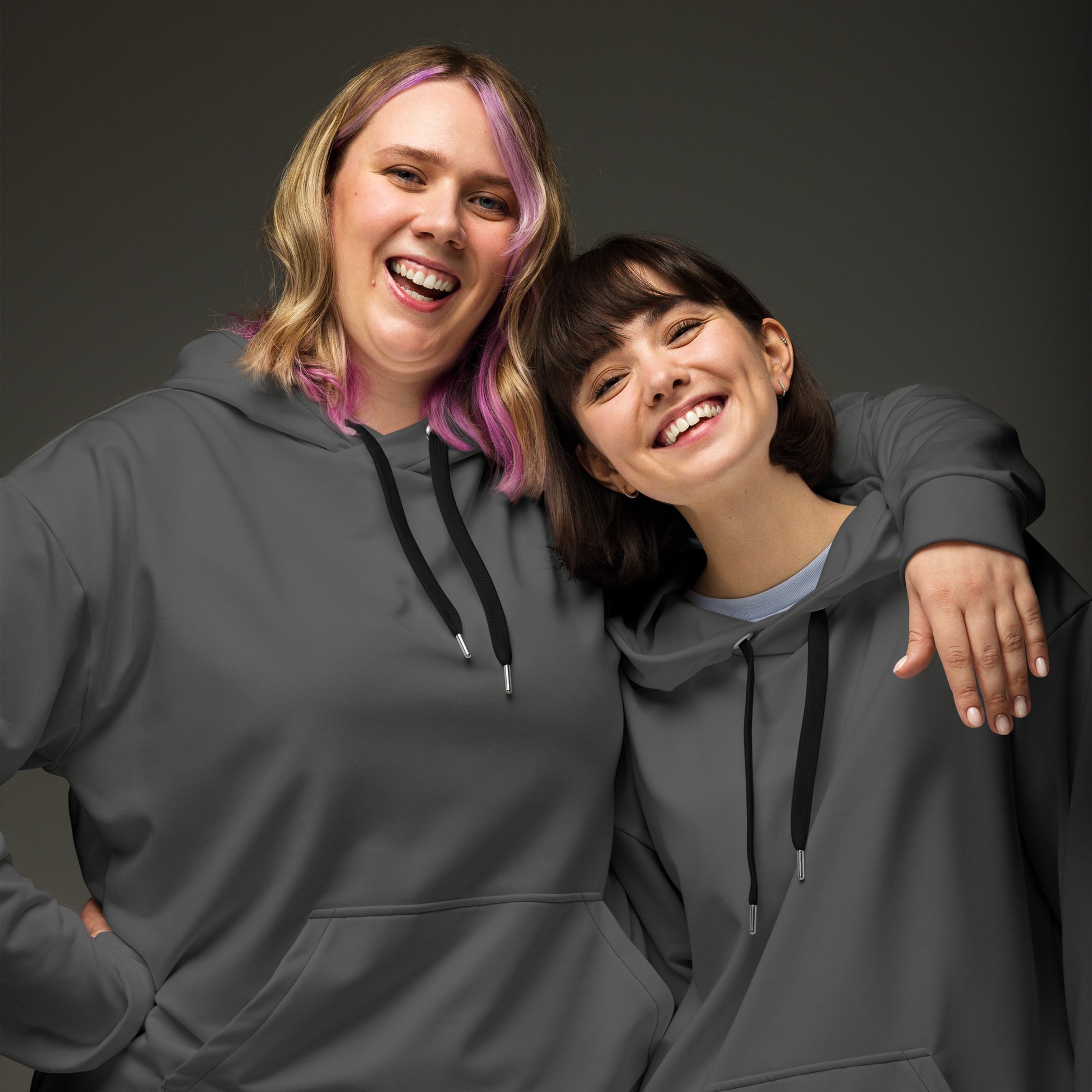 Two women smiling and posing together while wearing matching dark gray hoodies with black drawstrings, photographed against a dark gray studio background. Minimalist Comfortable Plain Gray Hoodie for Relaxed Wear