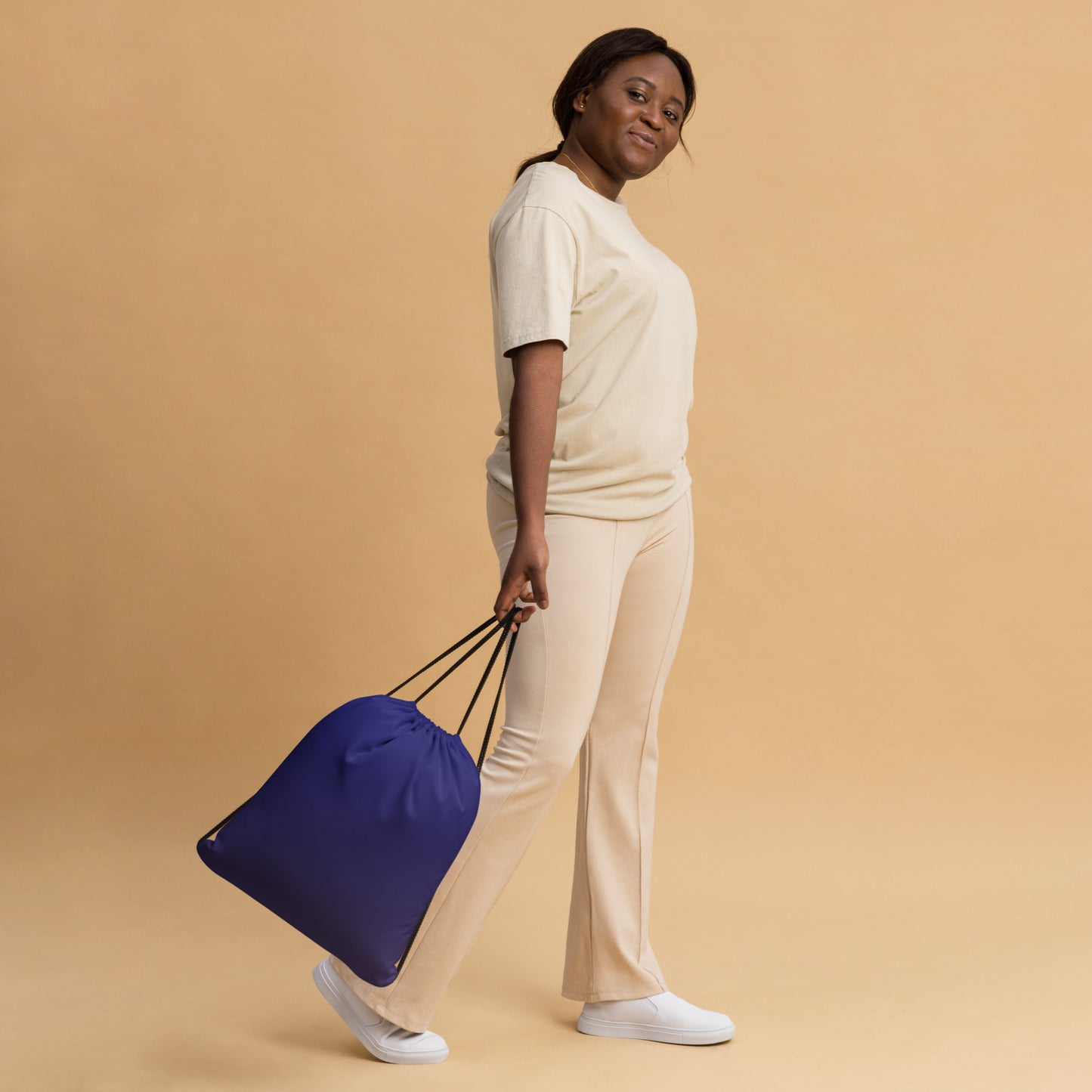 A woman in a cream-colored lounge set holds a navy blue drawstring bag with black handles against a solid beige studio background. Practical Bold Minimalist Solid Midnight Blue Drawstring Bag