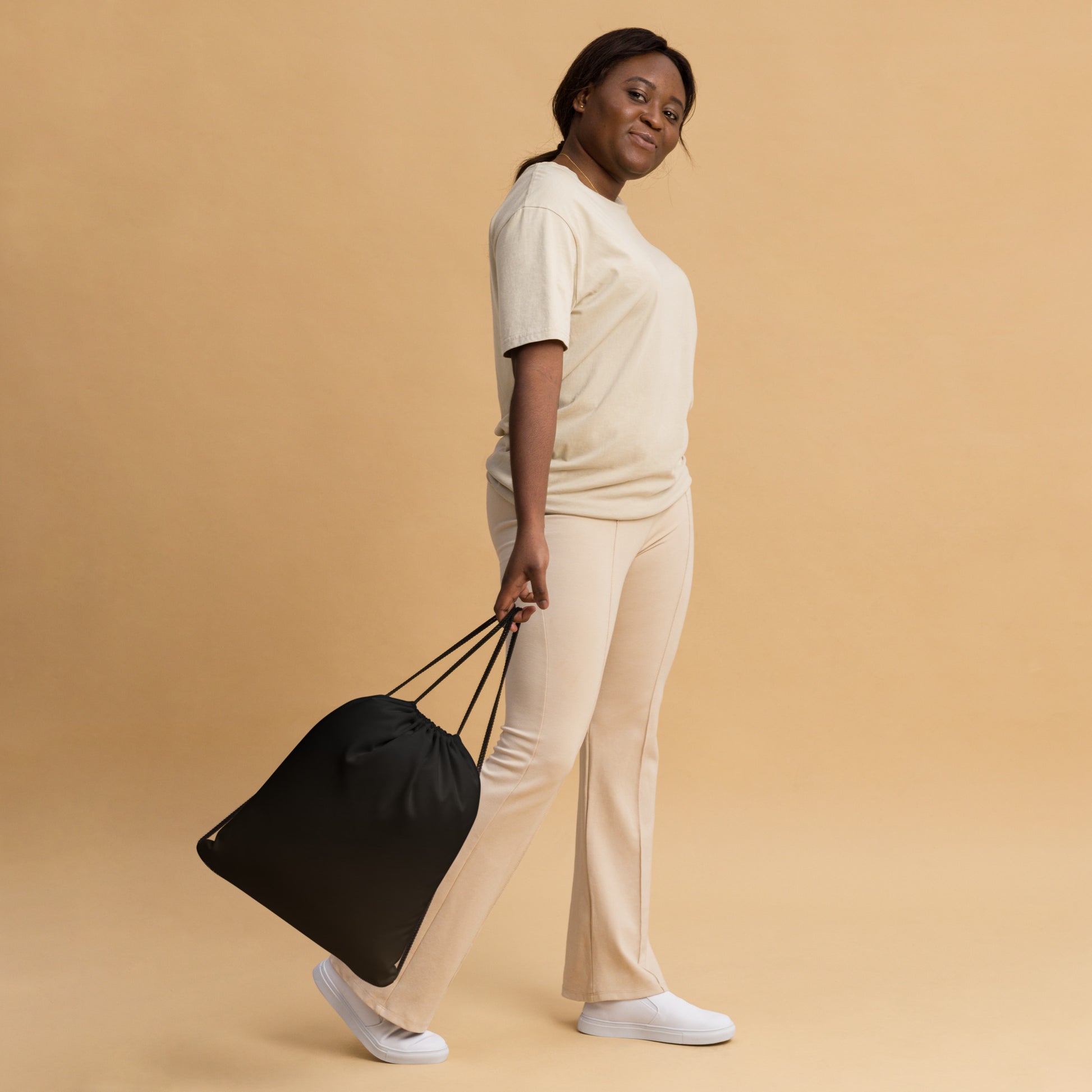 A woman in a cream-colored lounge set walks while carrying a black drawstring bag against a solid beige studio background. Simple Bold Minimalist Solid Black Lightweight Drawstring Bag