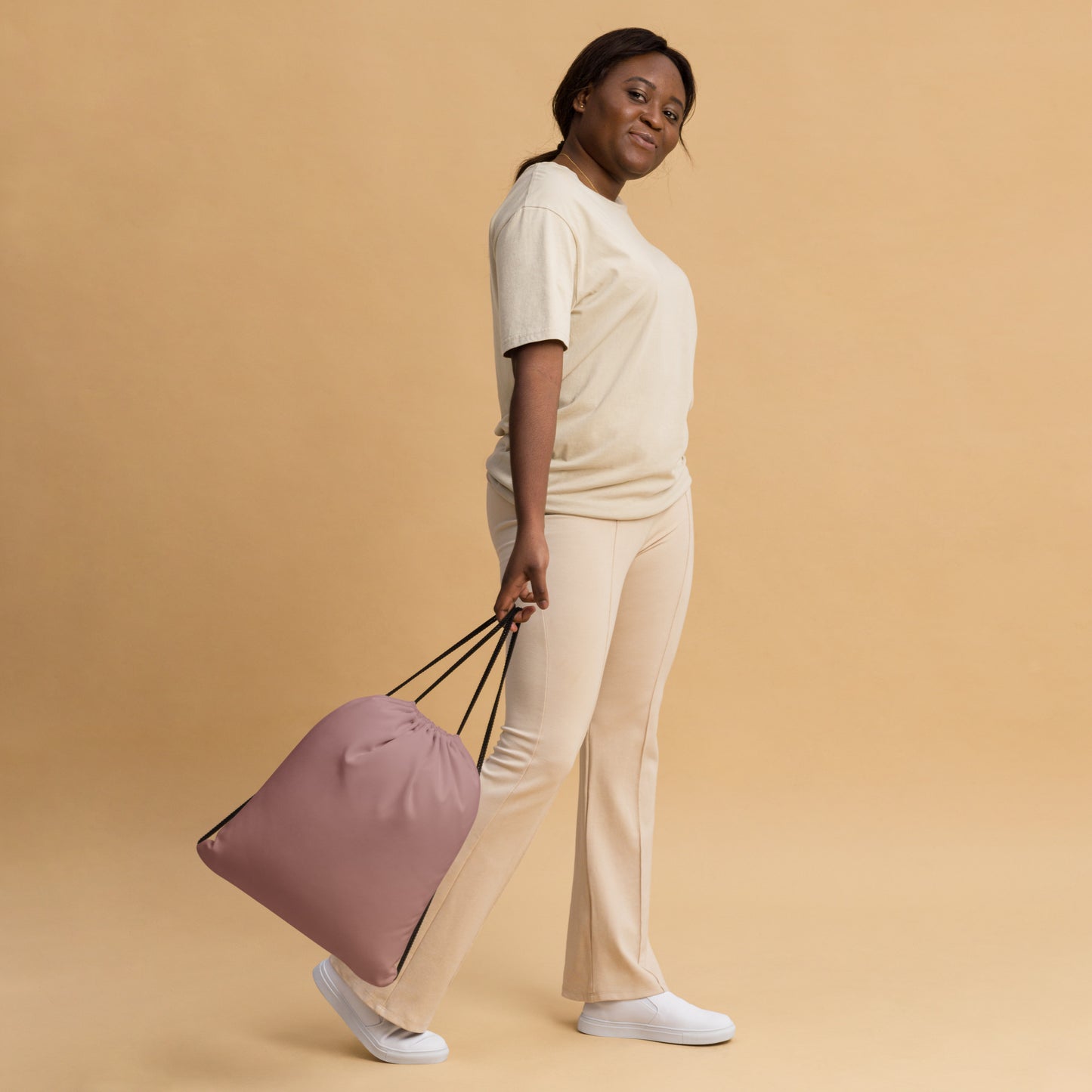 A woman in a cream-colored lounge set walks while carrying a dusty pink drawstring bag with black handles against a solid beige background. Stylish Minimalist Solid Rosy Brown Durable Drawstring Bag