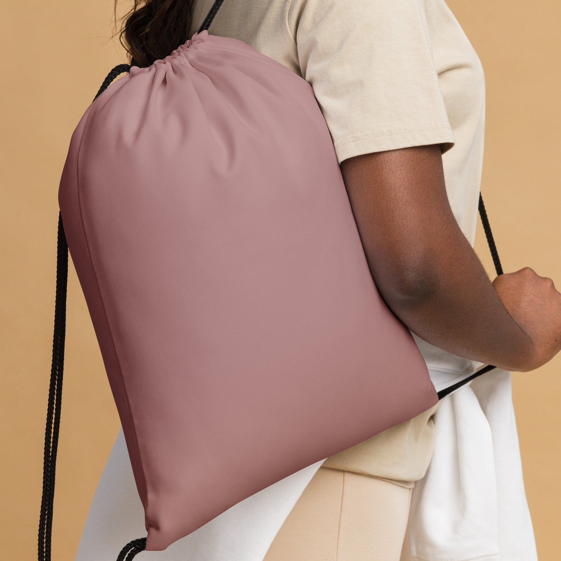 A person wearing a cream t-shirt and white shorts carries a dusty rose drawstring backpack against a solid beige background. Trendy Minimalist Solid Rosy Brown Durable Drawstring Bag